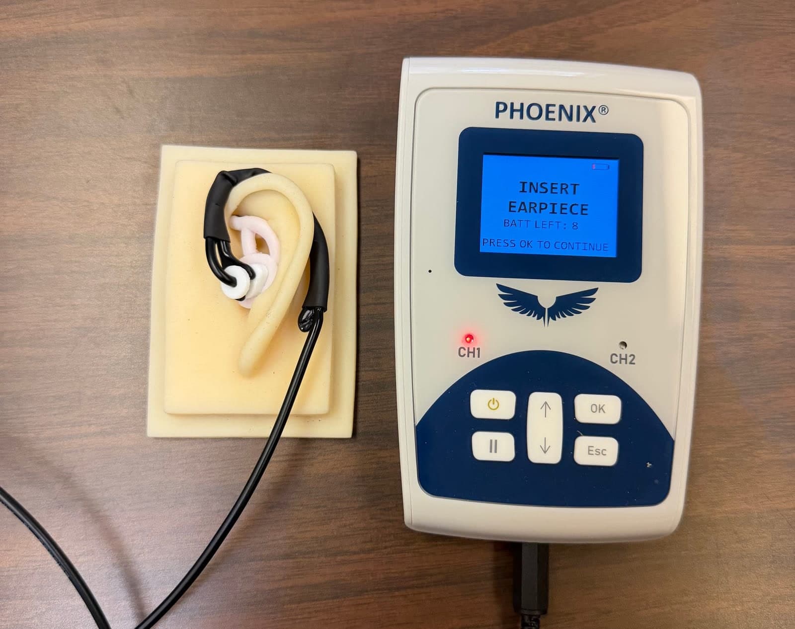 Evren's Phoenix controller next to an assembled ear-worn bioelectronic earpiece fitted to a silicone ear model, showing the CH1 channel active and prompting to insert the earpiece.